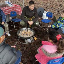 Students and one staff sitting around a gas firepit roasting marshmellows